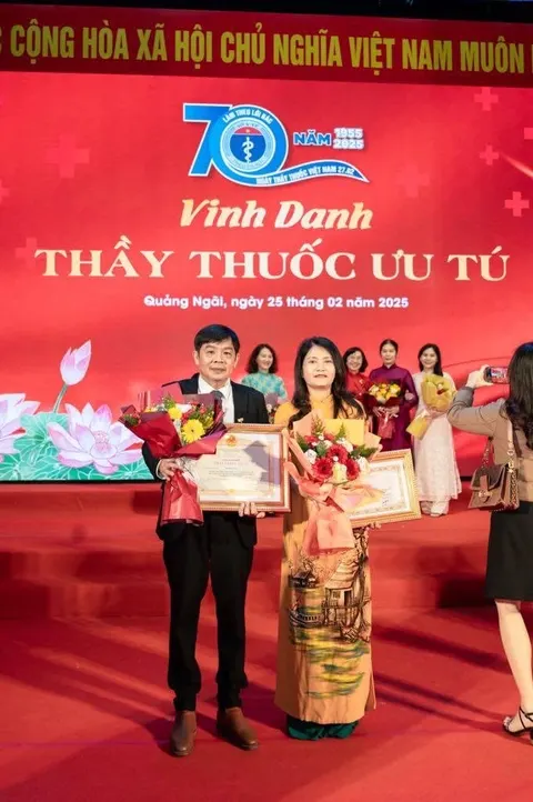 Bác sĩ Xuân Phi mặc áo dài và chồng mặc vest, cùng cầm giấy khen và hoa trên sân khấu Lễ kỷ niệm 70 năm ngày Thầy thuốc Việt Nam, phông nền đỏ ghi Vinh danh Thầy thuốc Ưu tú